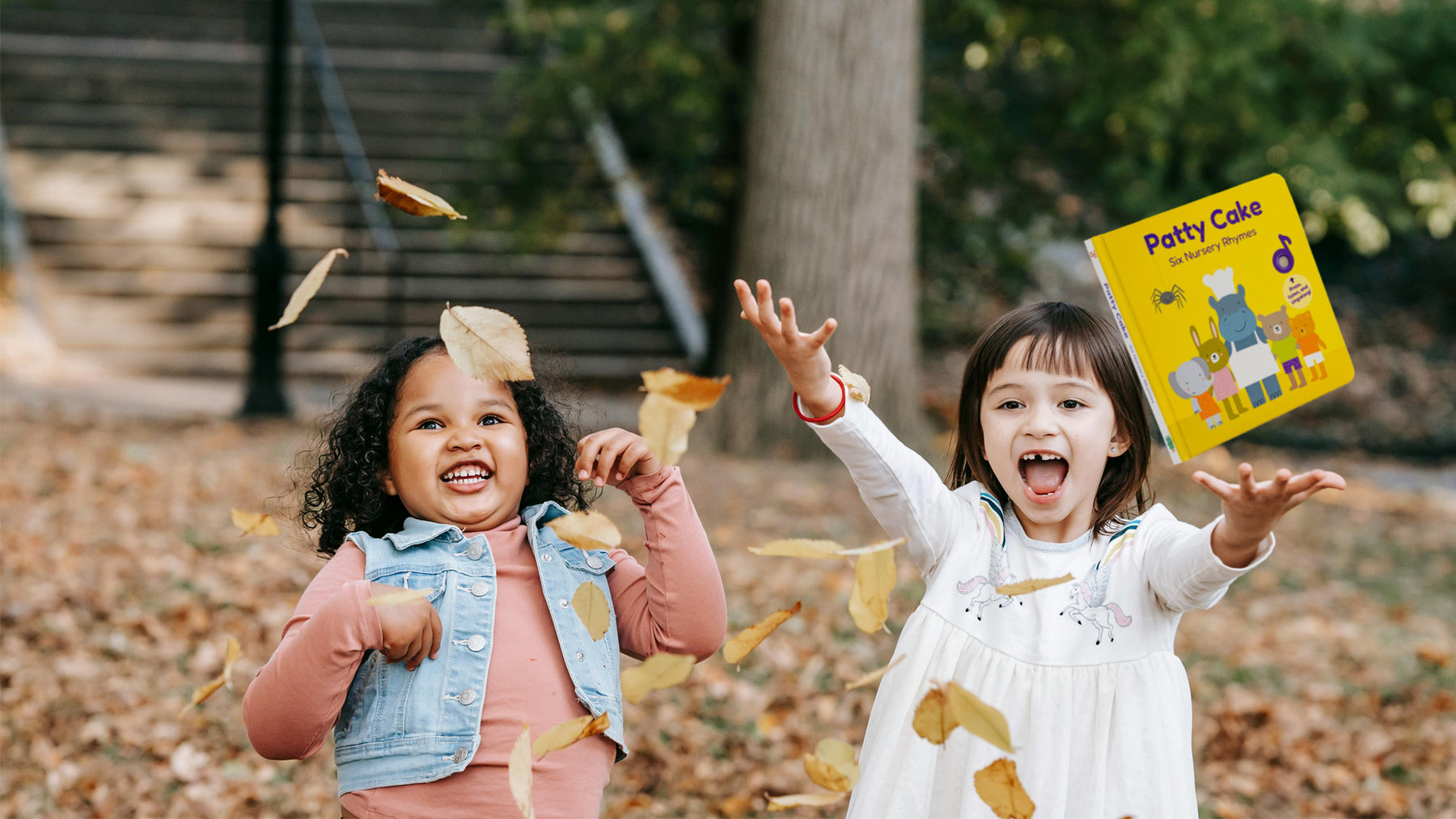 Two girls are throwing a picture book in some leaves in a park. Fall kids activities, toddler sensory activities, sensory activities, kids education.