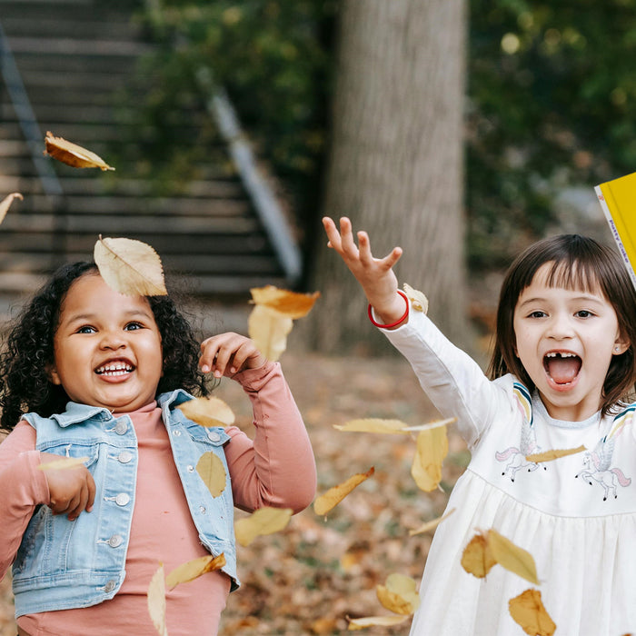 Two girls are throwing a picture book in some leaves in a park. Fall kids activities, toddler sensory activities, sensory activities, kids education.