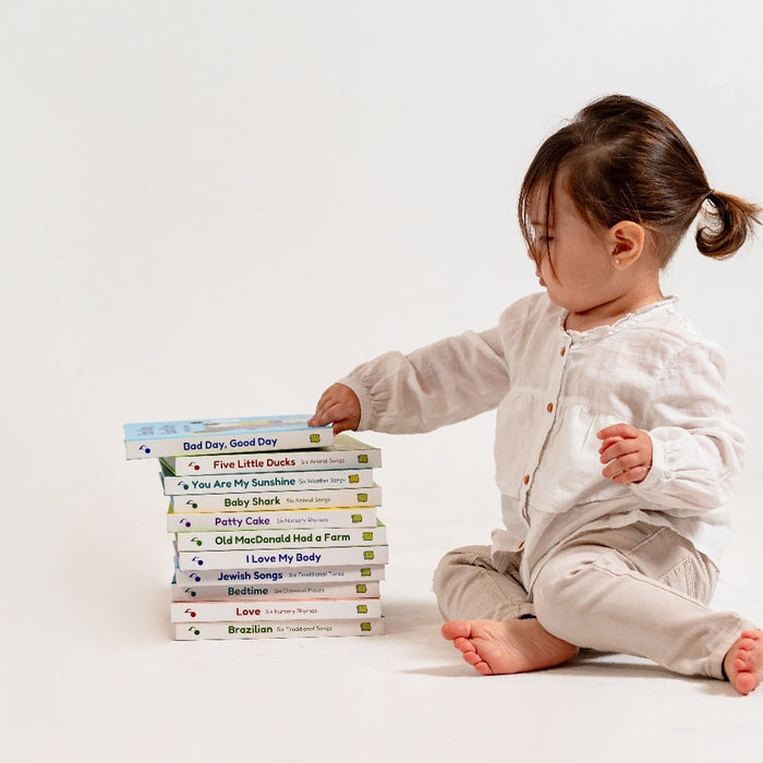 A toddler girl is touching a pile of sound books. Picture books, musical books, kids books, bedtime books.